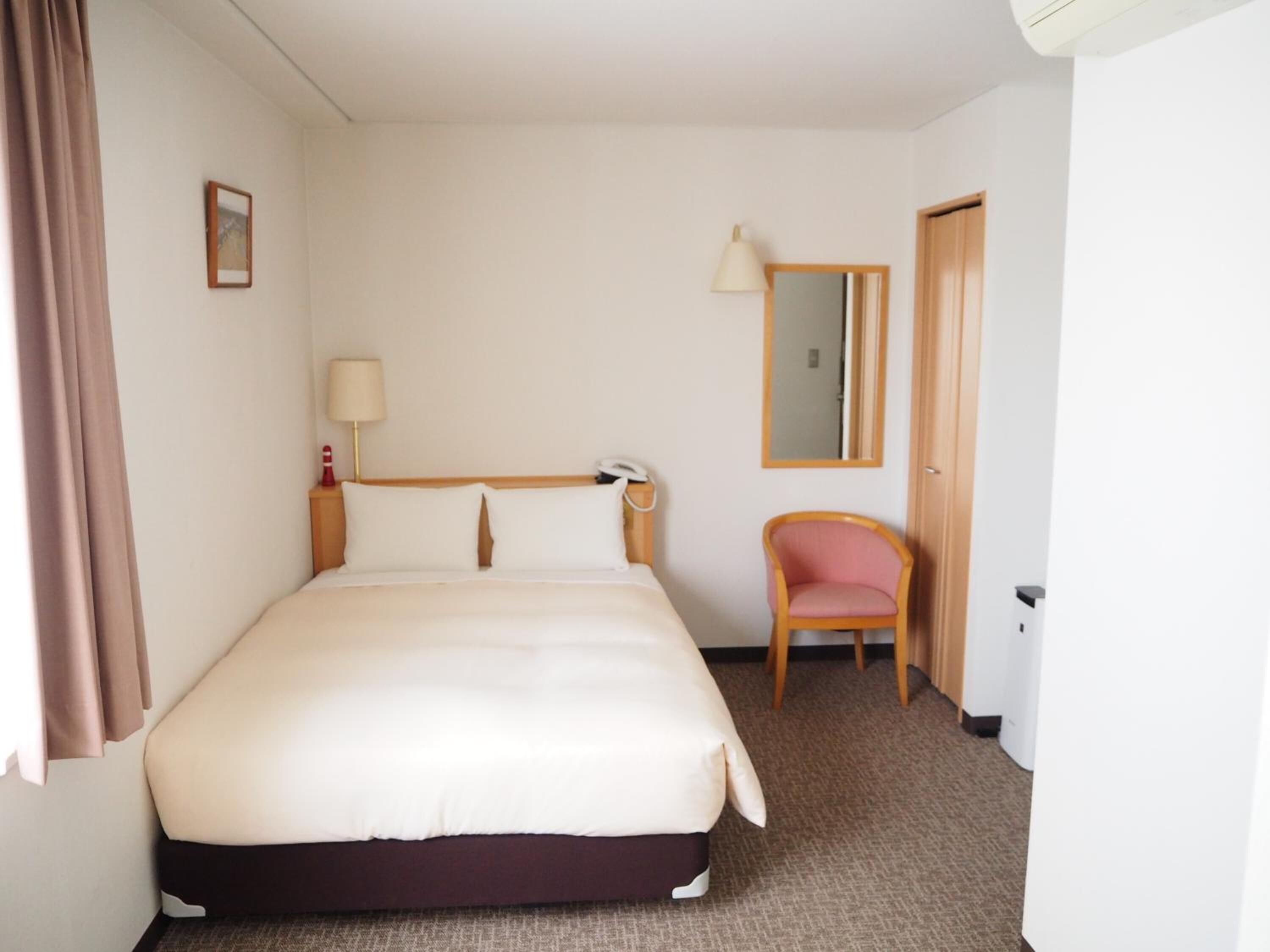 Een hotelkamer met een bed, een stoel, een bureau, een lamp, een spiegel en een deur.