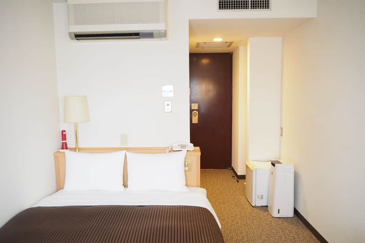A hotel room with a bed, a lamp, a telephone, an air conditioning unit, and a door leading to another room.