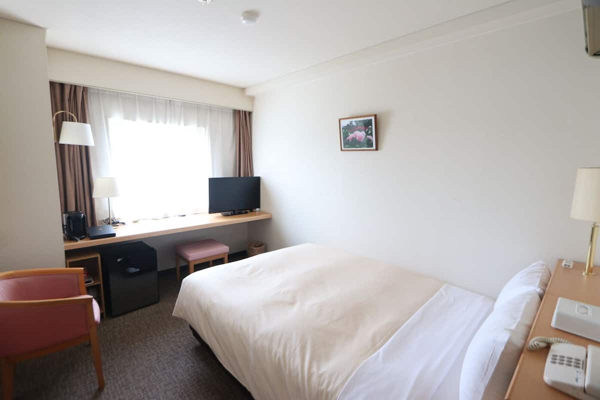 A hotel room with a bed, desk, chair, and a small window with curtains.