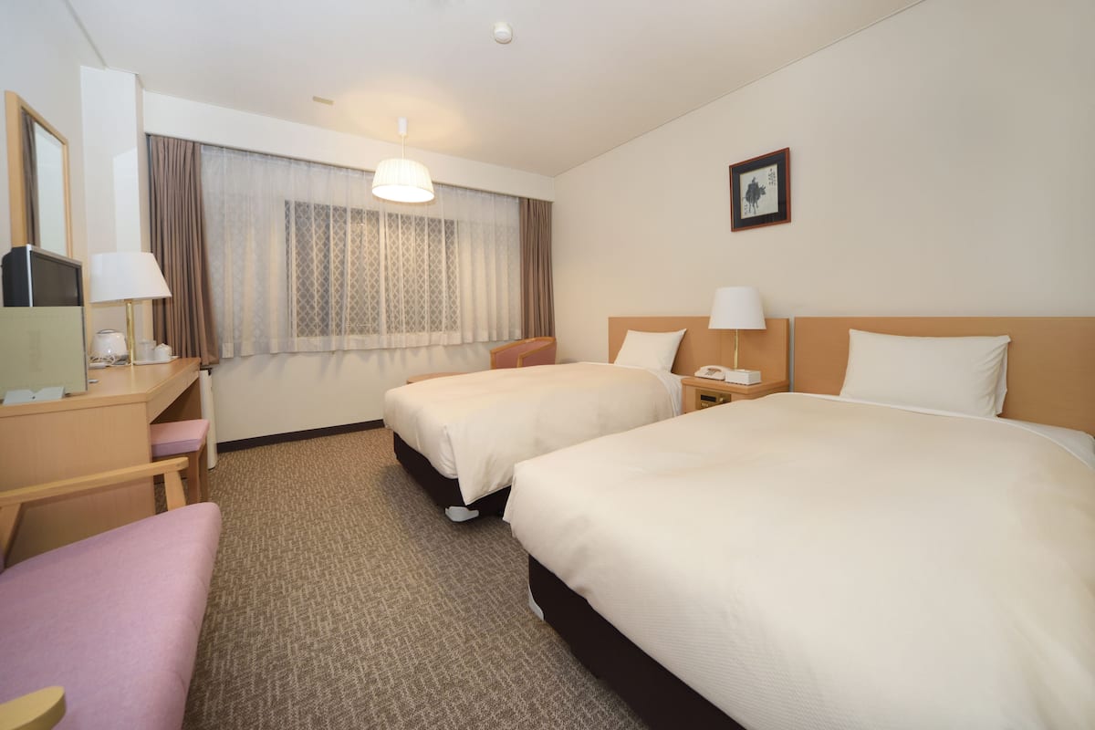 A hotel room with two beds, a desk, a chair, a lamp, and a window with curtains.