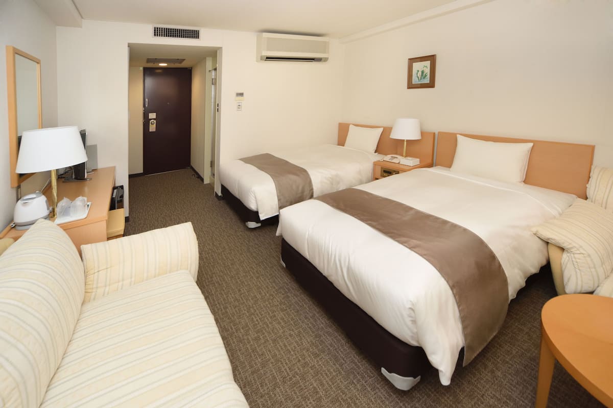 A hotel room with two beds, a desk, a chair, a lamp, and an air conditioning unit.