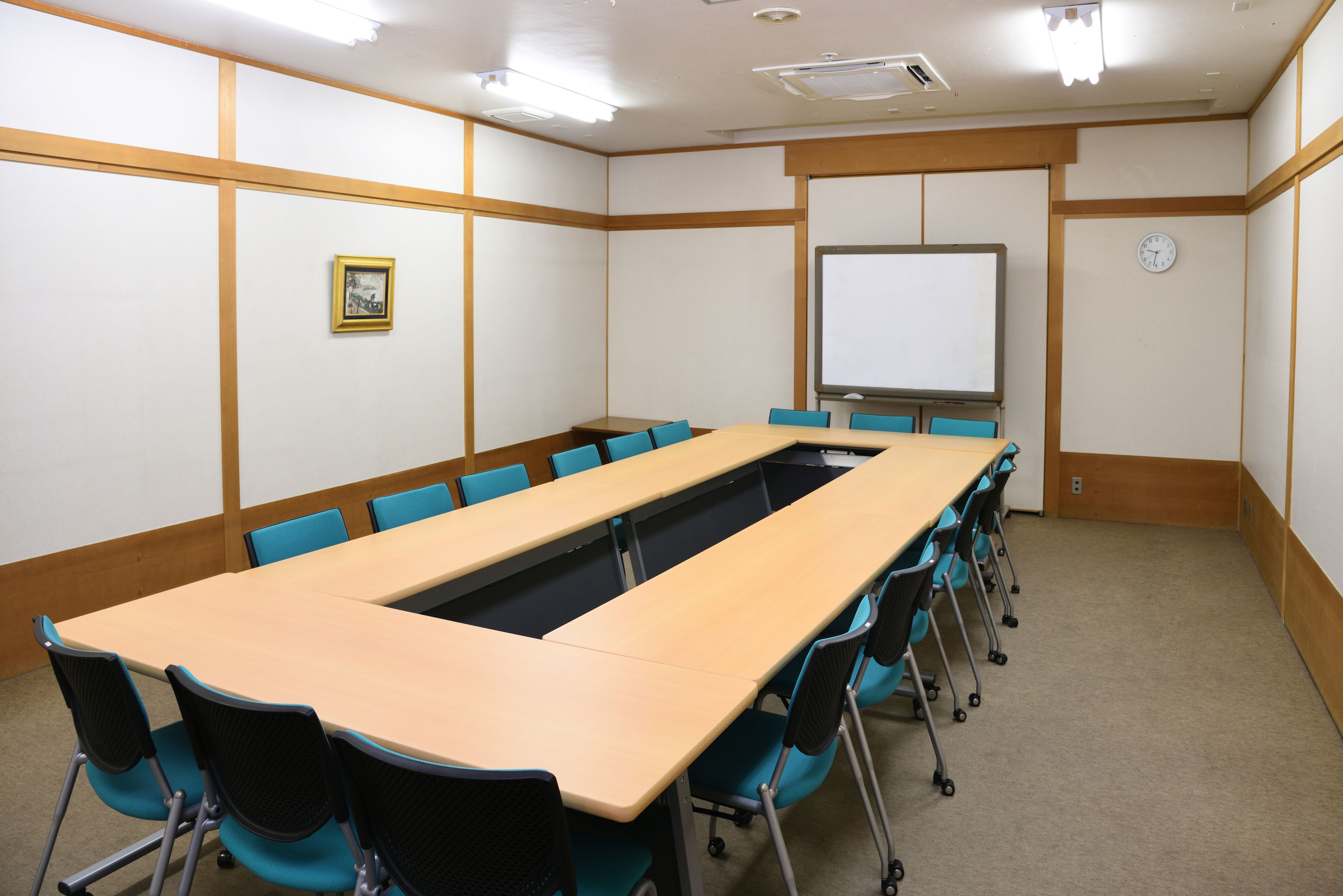 A conference room with a long table, blue chairs, a whiteboard, and a clock on the wall.