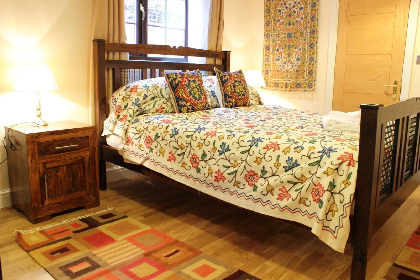 A bed with a floral bedspread, a wooden nightstand with a lamp, and a patterned rug on a wooden floor.