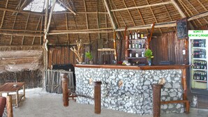 A rustic bar area with a stone counter, wooden stools, and a thatched roof.