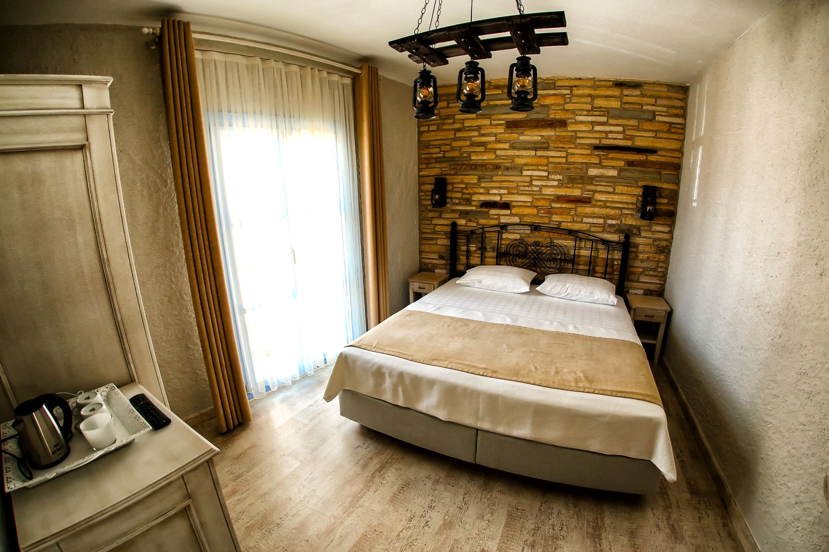 A bedroom with a stone wall, a bed with a beige bedspread, a nightstand with a lamp, and a window with curtains.