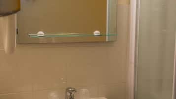 Une salle de bain avec un lavabo blanc, une étagère en verre où sont rangés les articles de toilette, un miroir et un espace douche.