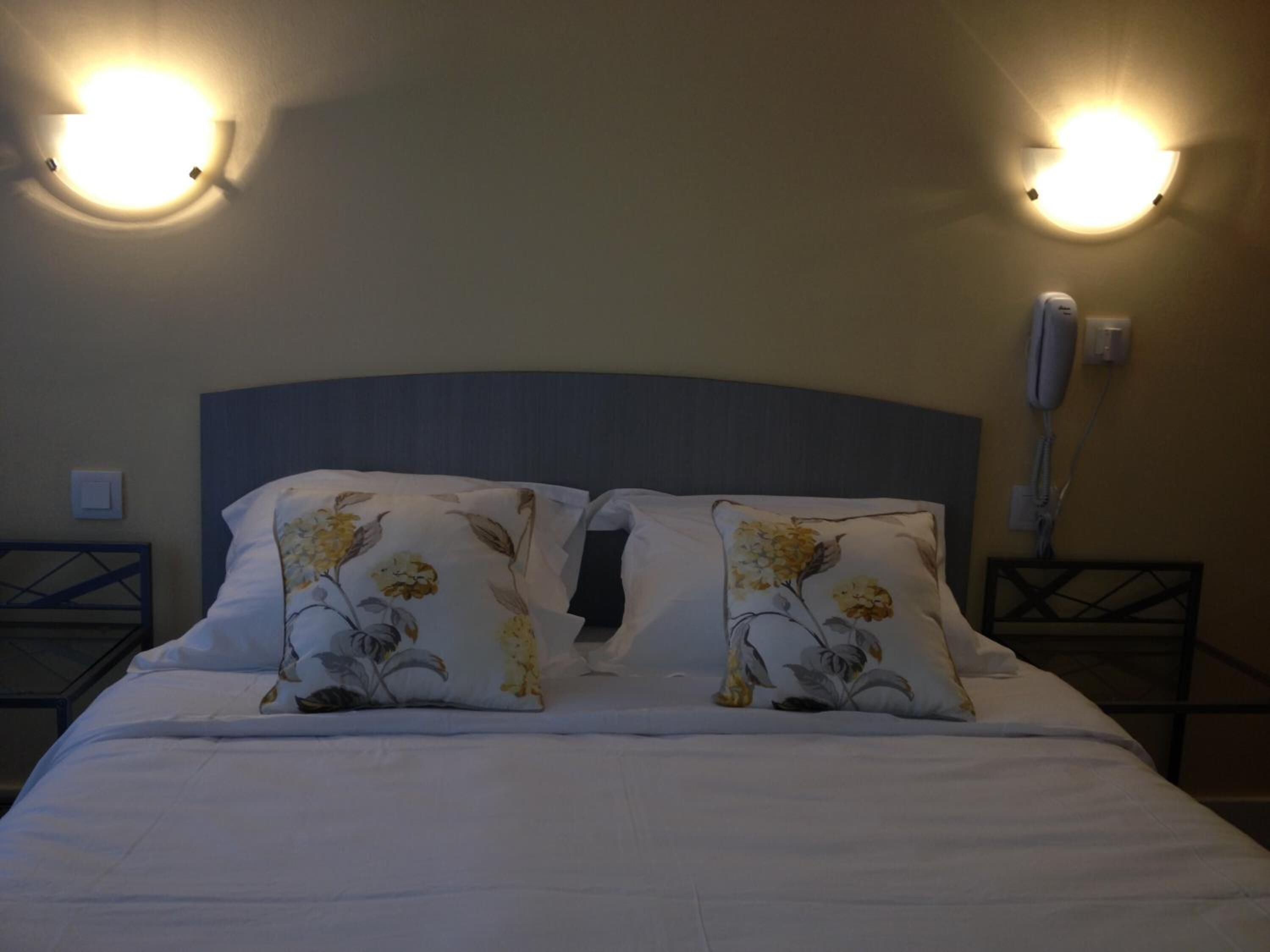 Una cama bien hecha con almohadas estampadas, un cabecero oscuro y dos lámparas fijadas a la pared.