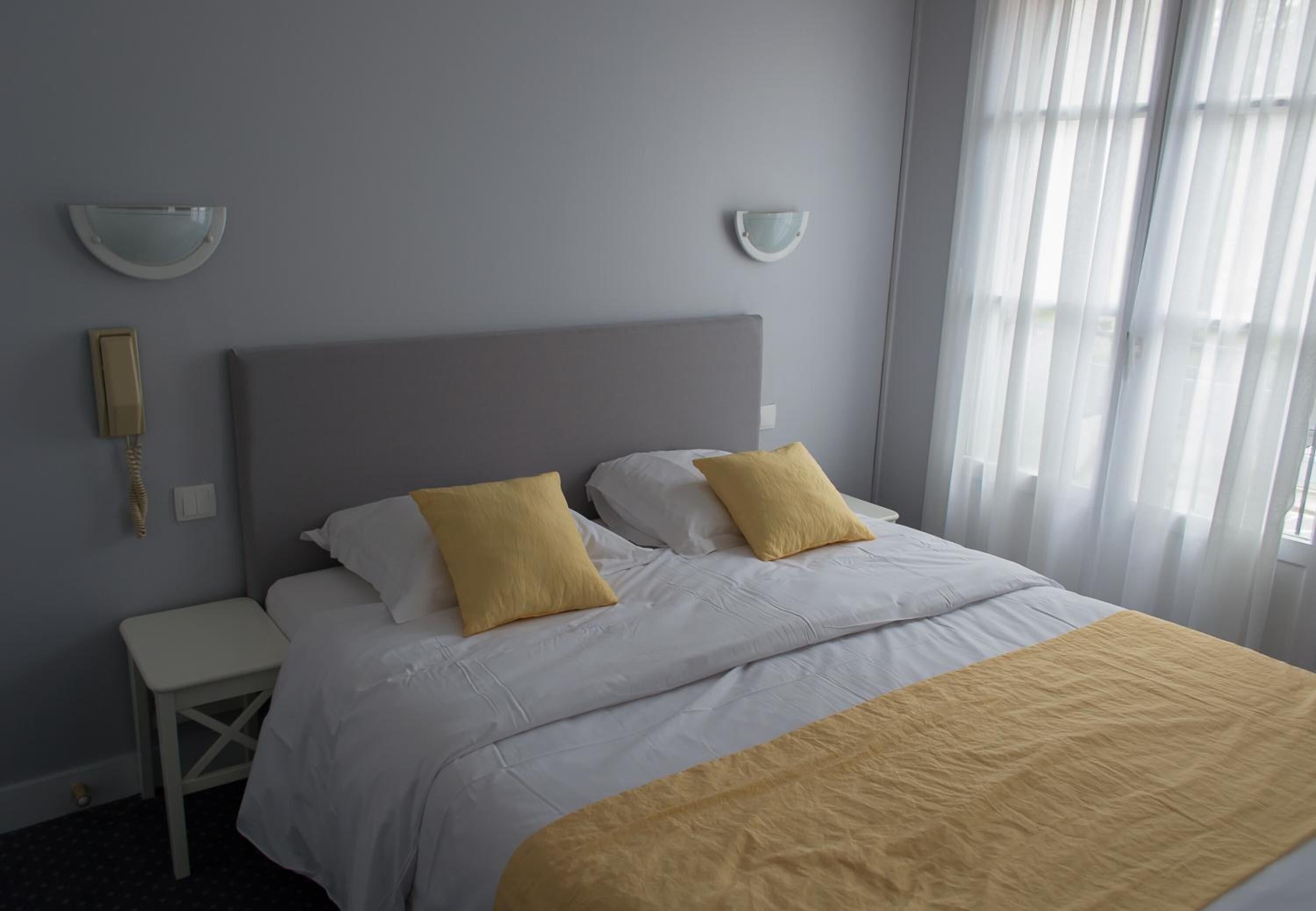 Una cama bien hecha con ropa de cama blanca y amarilla, una mesita de noche y una lámpara de pared.