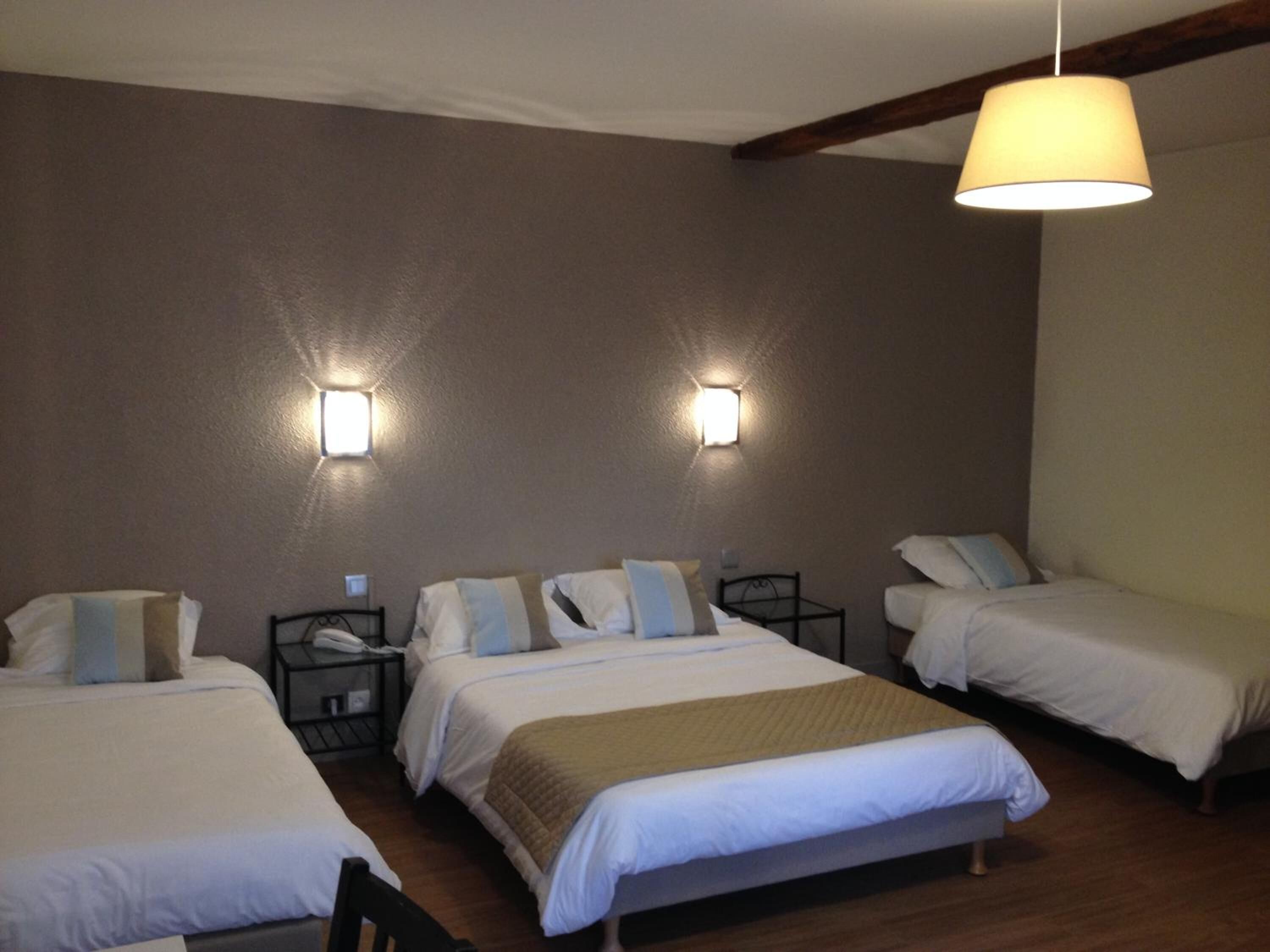 Une chambre d’hôtel avec trois lits, chacun doté de draps blancs et d’un plaid marron. On y trouve deux appliques murales et un luminaire suspendu.