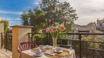 A table set for two with a vase of flowers, a teapot, cups, and a placemat on a balcony overlooking a residential area.