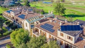 Um edifício residencial com vários painéis solares no telhado, rodeado por vegetação exuberante e um campo de golfe ao fundo.