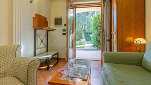 Quarto com sofá, mesa de centro com revistas, mesa lateral e porta que dá para um jardim.