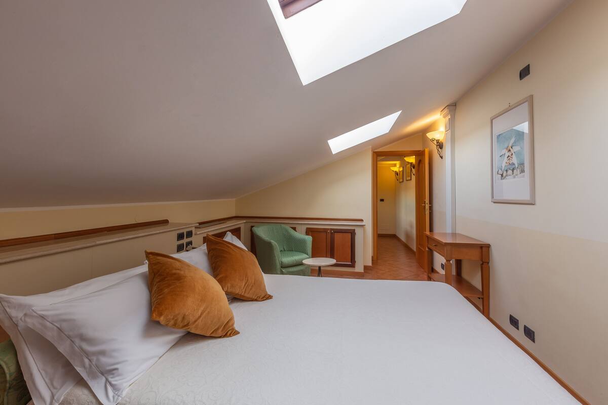 A bedroom with a skylight, a bed with pillows, a chair, a small table, and a painting on the wall.