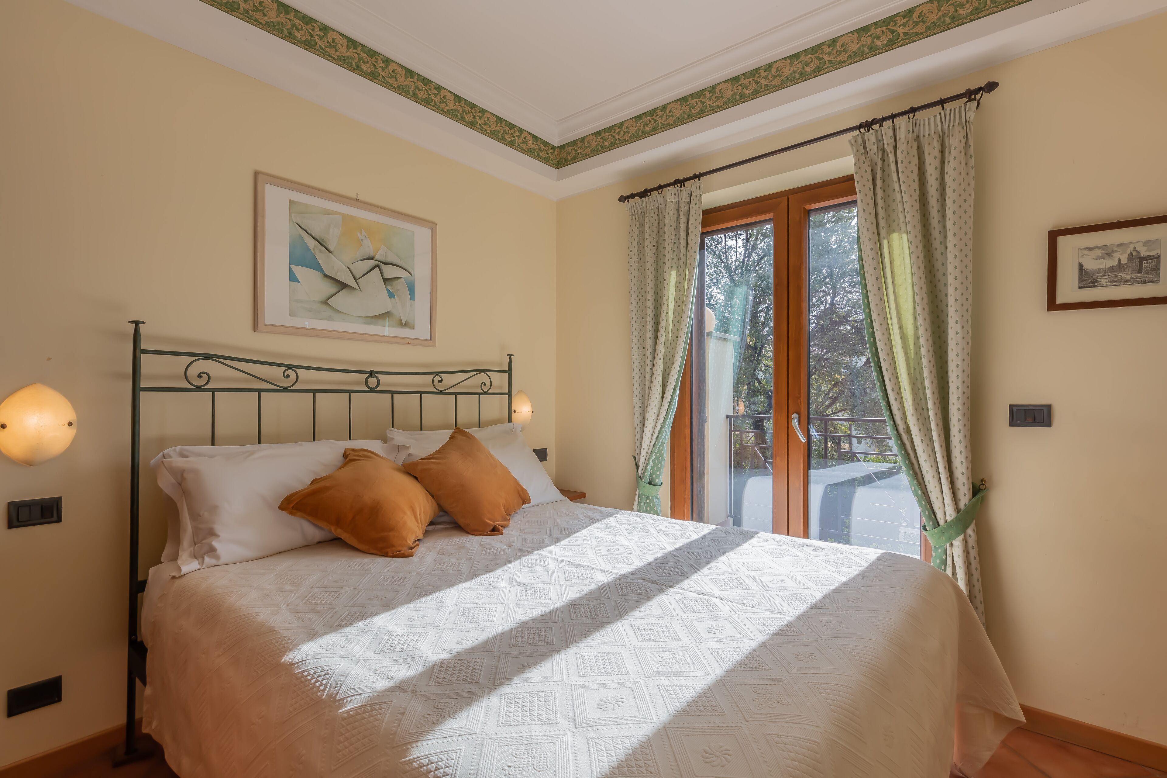 A neatly made bed with a patterned bedspread, a framed abstract painting above it, and a view of the outdoors through a window with curtains.