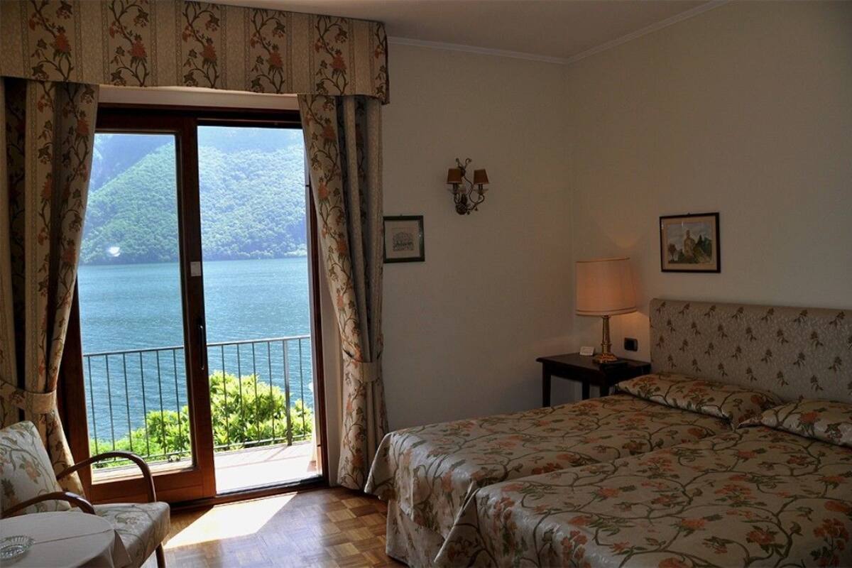 A hotel room with a bed, a small table, a lamp, and a view of a lake through a sliding door.