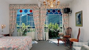 Habitación de hotel con cama, escritorio, silla y vistas a la vegetación a través de amplias ventanas con cortinas.