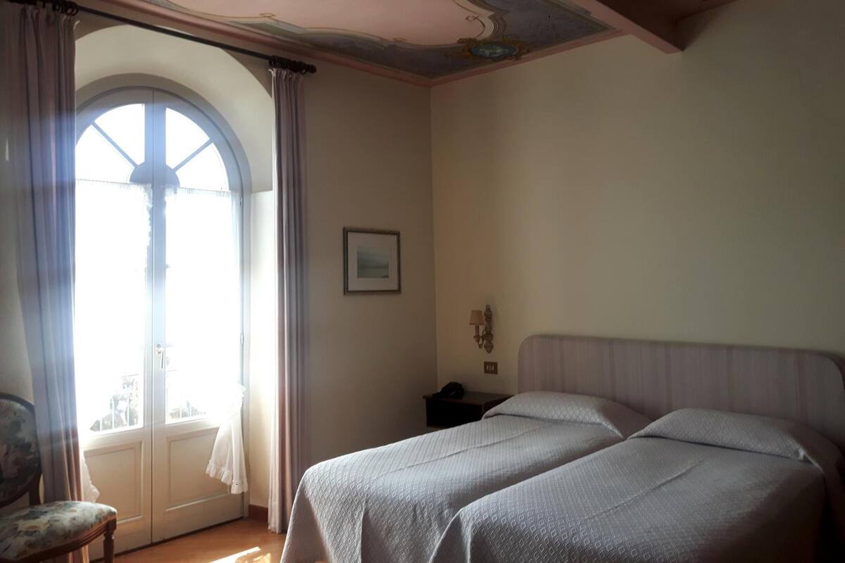A bedroom with two beds, a window with curtains, a framed picture on the wall, and a small table with a lamp.