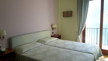 Quarto com duas camas, mesa de cabeceira, abajur, pintura e vista para o mar através de uma varanda.