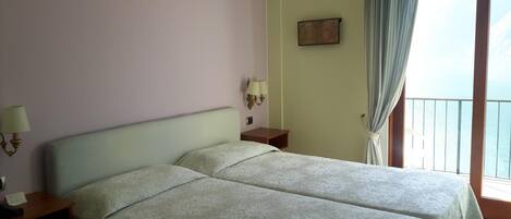 A room with two beds, a nightstand, a lamp, a painting, and a view of the sea through a balcony.