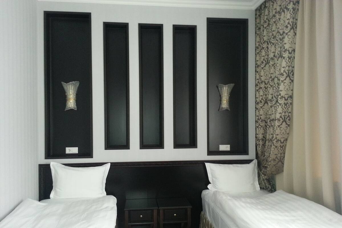 A hotel room with two single beds, a dark headboard, a small table, and wall-mounted sconces.