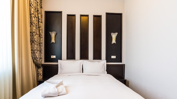 A neatly made bed with white linens and a folded towel, a dark headboard with vertical panels, and a wall-mounted decorative piece. - Royal Petrol Hotel Taldykorgan (Taldykorgan)