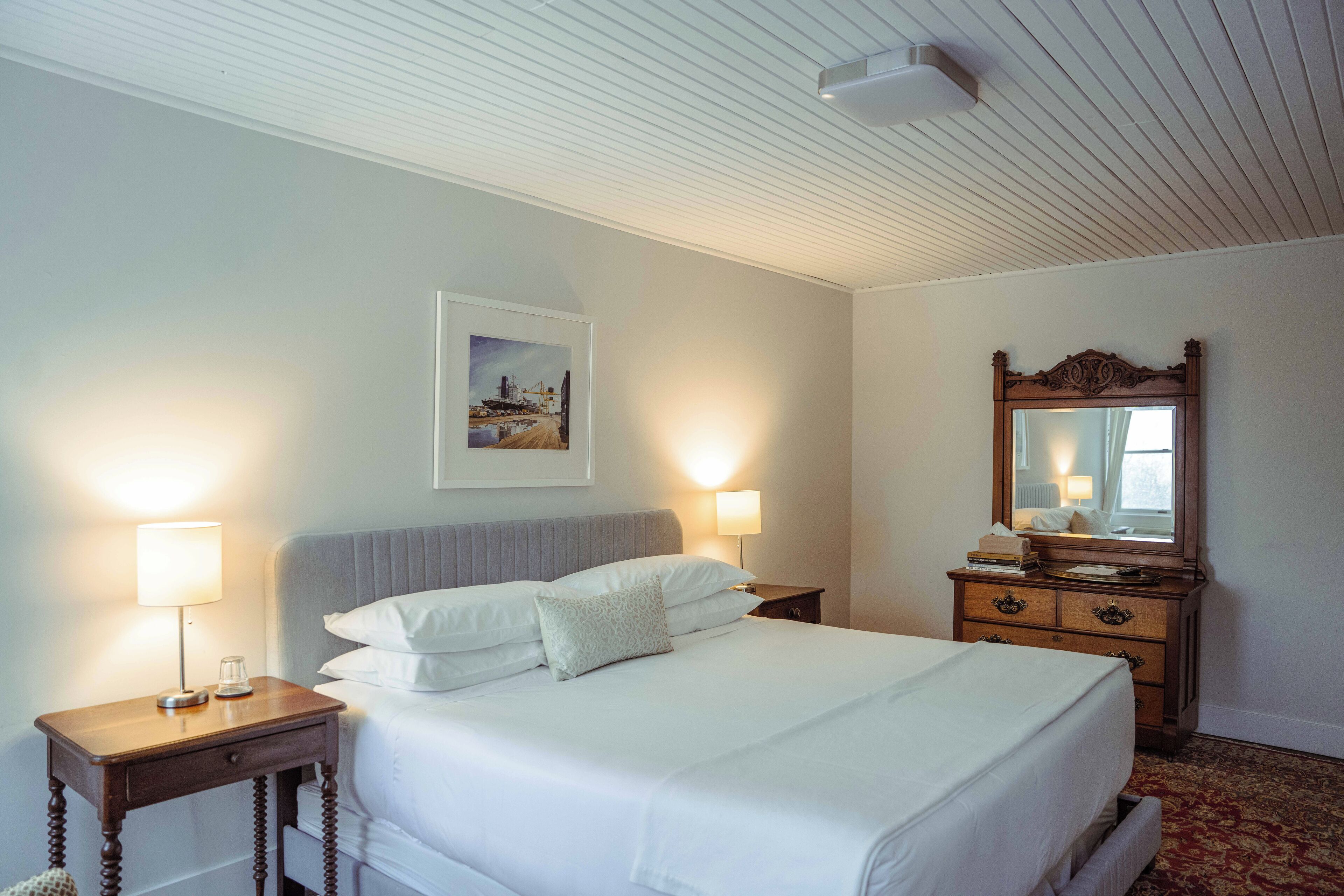 A neatly made bed with white linens, two bedside lamps, a framed picture on the wall, a wooden dresser with a mirror, and a patterned carpet.