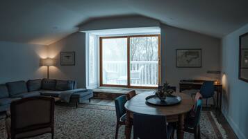 Uma sala de estar acolhedora com um sofá, uma mesa de jantar com cadeiras, um abajur e uma grande janela com vista para uma varanda.