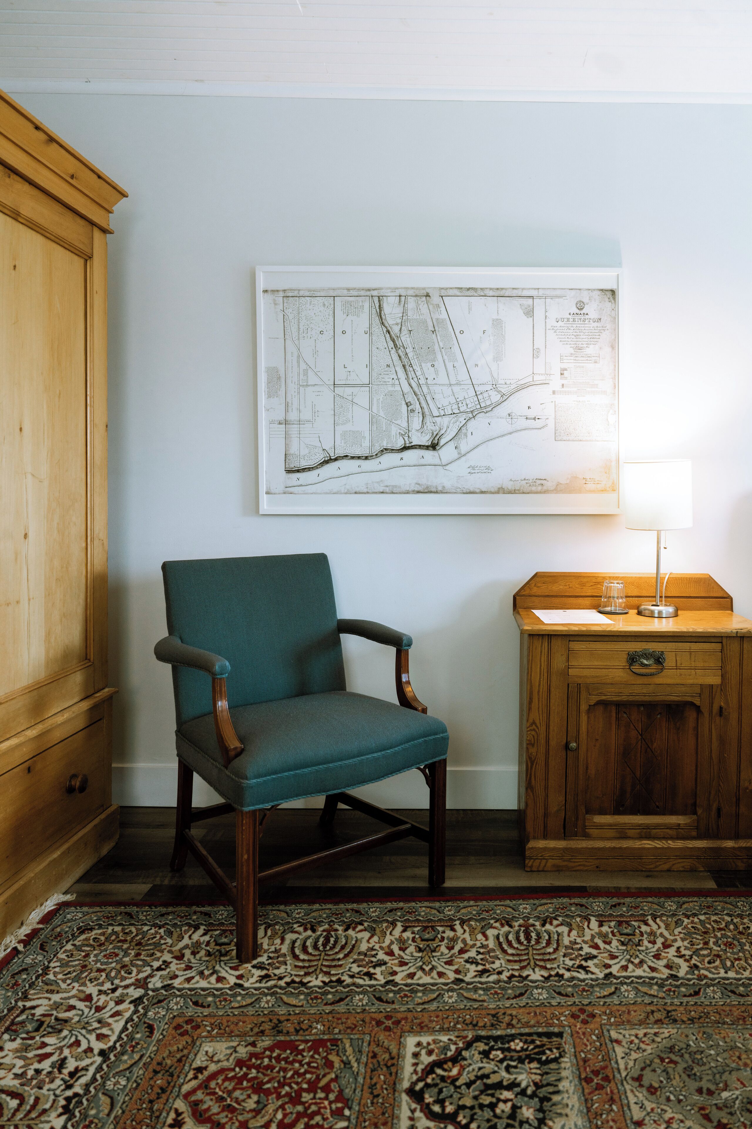 A room with a wooden cabinet, a teal armchair, a wooden side table with a lamp, and a large map on the wall.