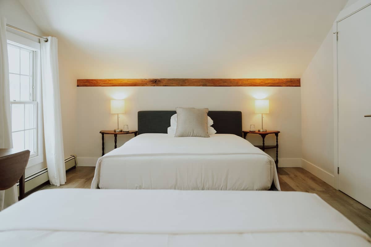 Une chambre avec deux lits, une poutre en bois au plafond, des lampes de chevet et une fenêtre avec des rideaux.