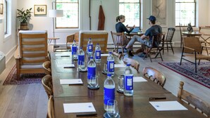 Una sala de conferencias con una larga mesa, sillas, botellas de agua y una persona usando un ordenador portátil.