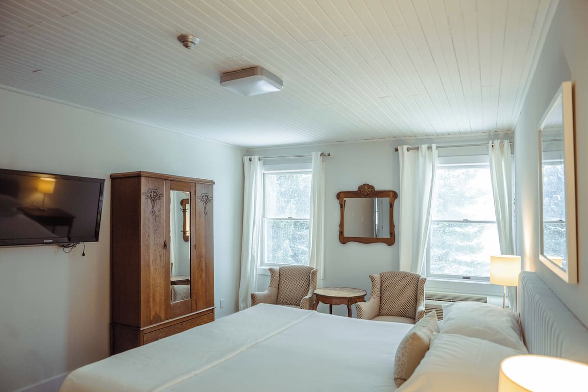 Une chambre d’hôtel avec un grand lit, une armoire en bois, une télévision, une petite table, deux chaises et une fenêtre avec des rideaux.