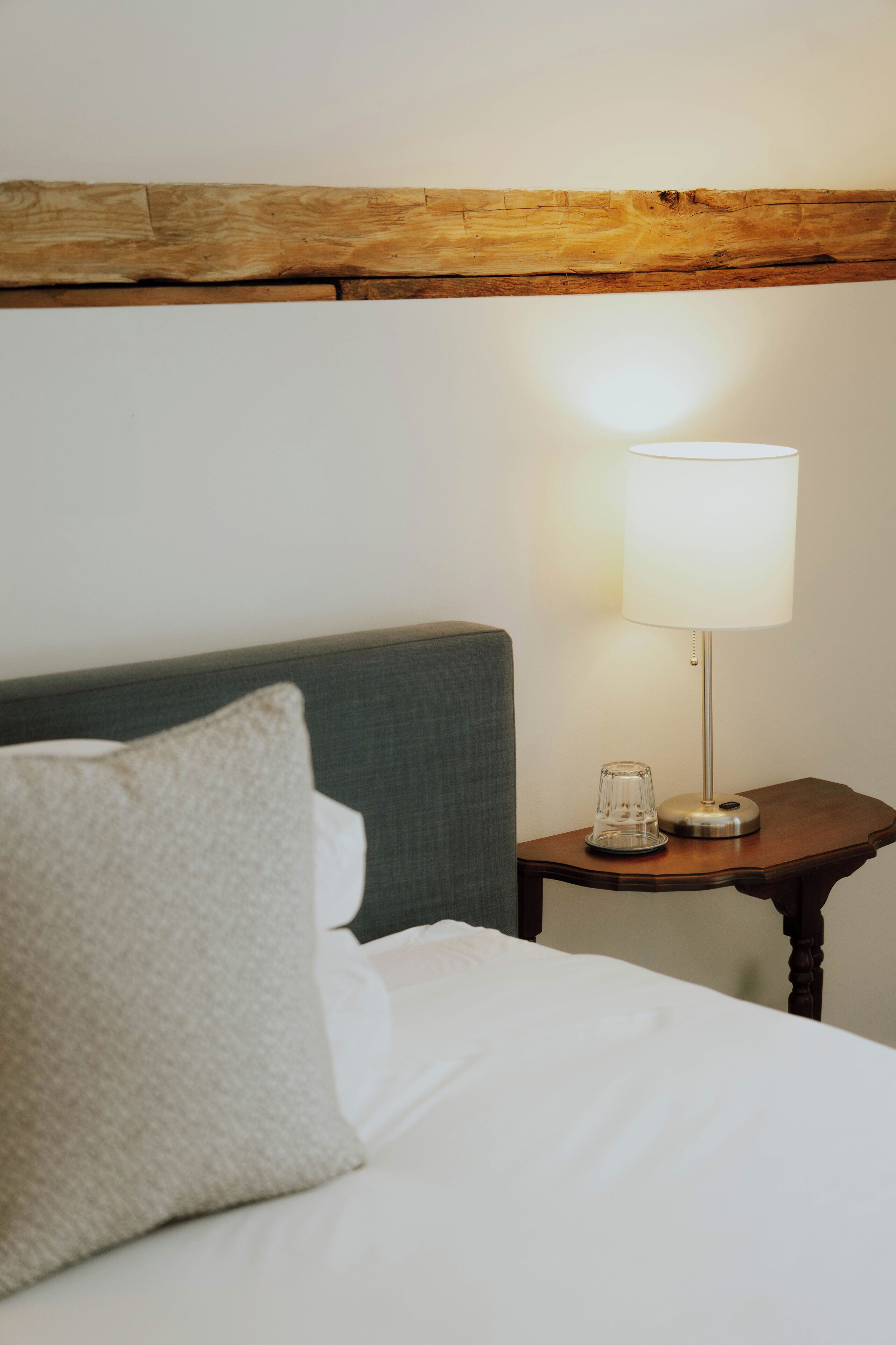 Una cama bien hecha con sábanas blancas y almohada gris, una mesita de noche con lámpara y cabecera de madera.