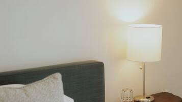 Un letto rifatto con lenzuola bianche e un cuscino grigio, un comodino con una lampada e una testiera in legno.