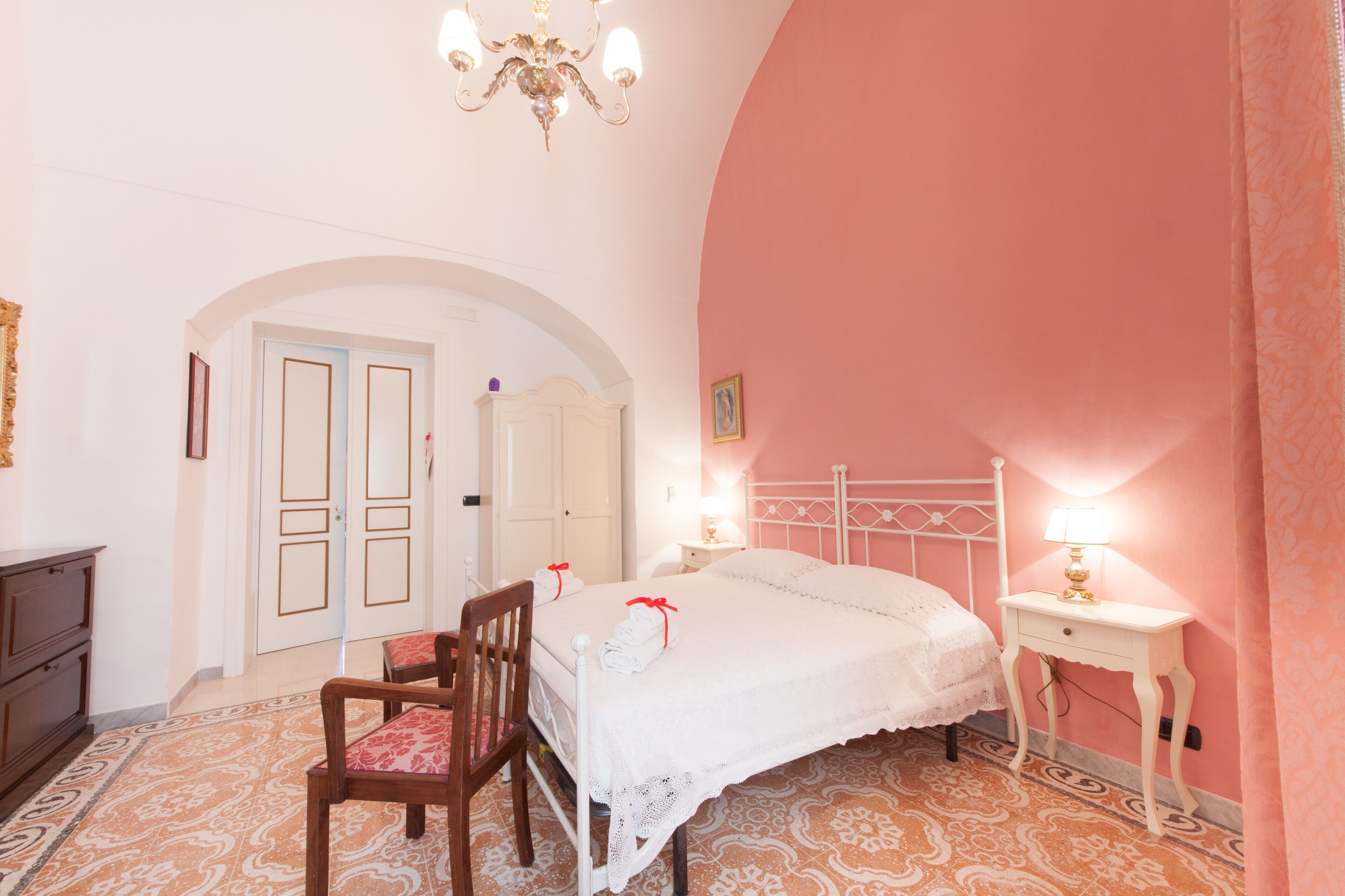 A bedroom with a white bed, a wooden chair, a bedside table with a lamp, and a chandelier.