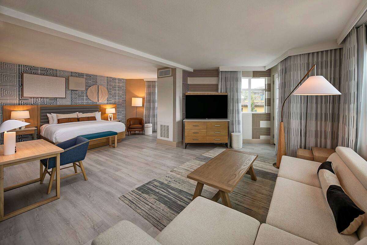 A modern hotel room with a large bed, a TV, a sofa, a coffee table, and a desk with a lamp.