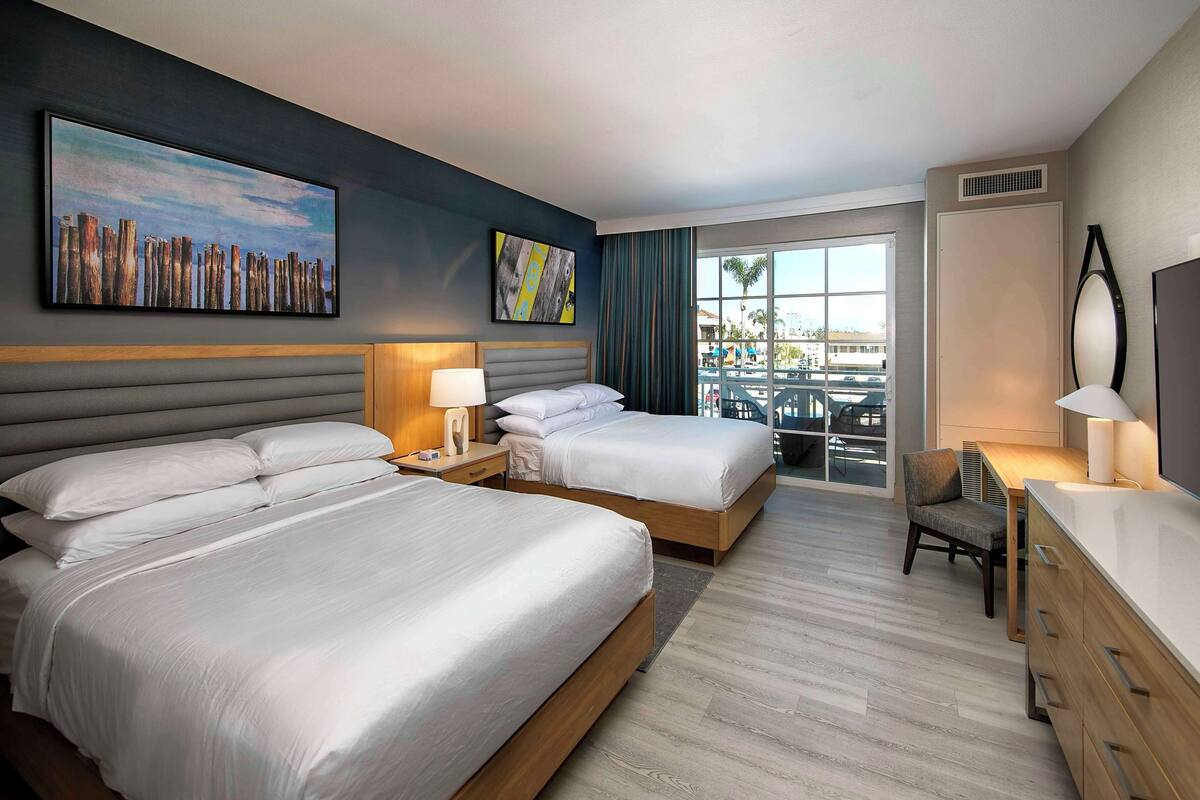 A hotel room with two beds, a desk, a chair, a TV, and a balcony with a view of palm trees and buildings.