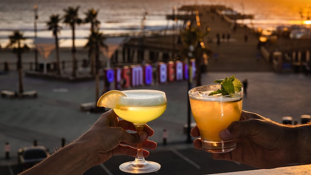 Two cocktails being held by hands against a sunset beach backdrop. - Inn at the Pier Pismo Beach, Curio Collection by Hilton (Pismo Beach)