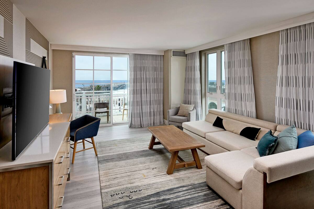 A modern living room with a sofa, coffee table, and TV. Large windows with curtains provide a view of the city.