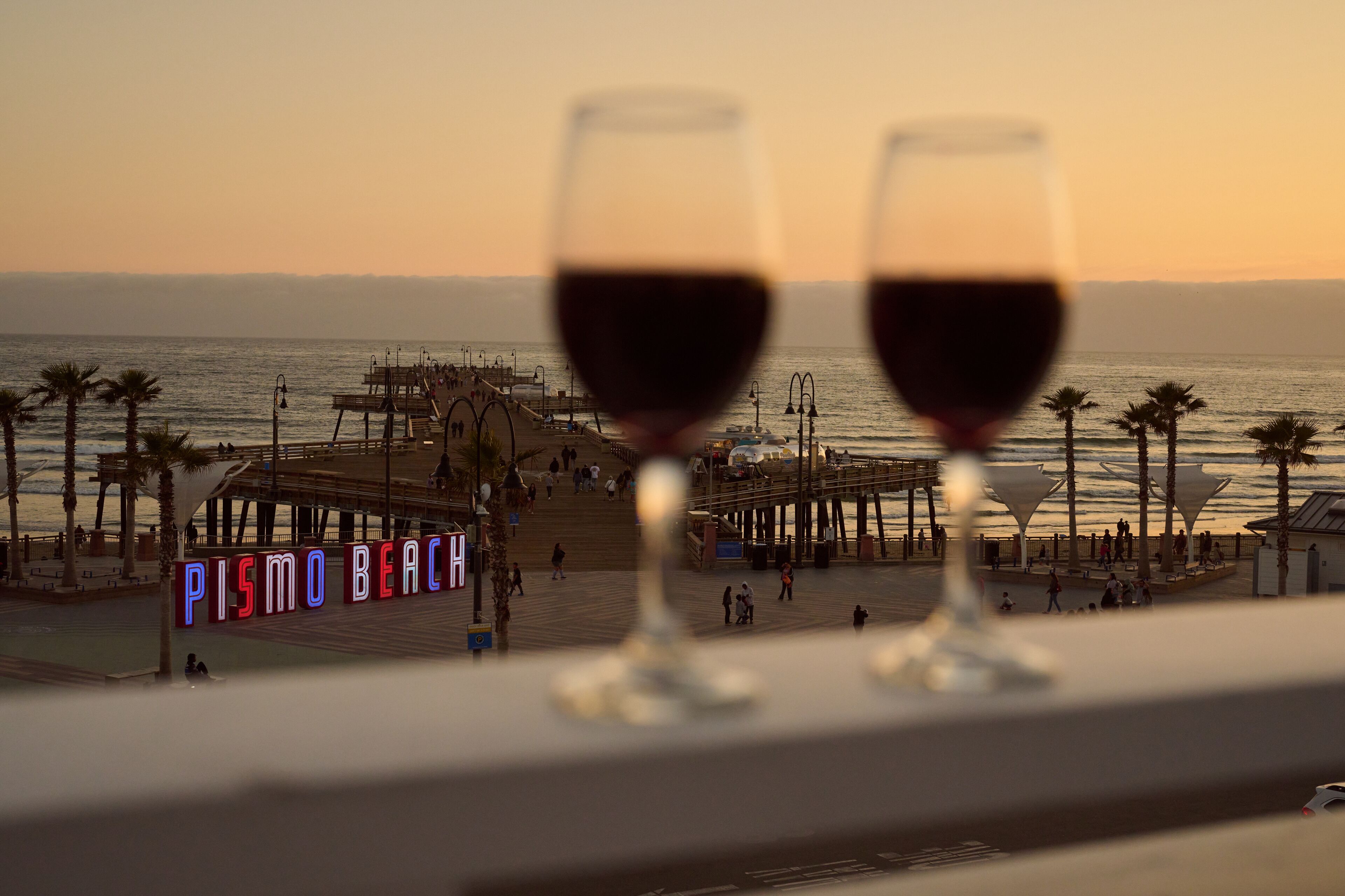 Duas taças de vinho numa varanda com vista para a Praia de Pismo, onde se vê um cais e palmeiras ao fundo.