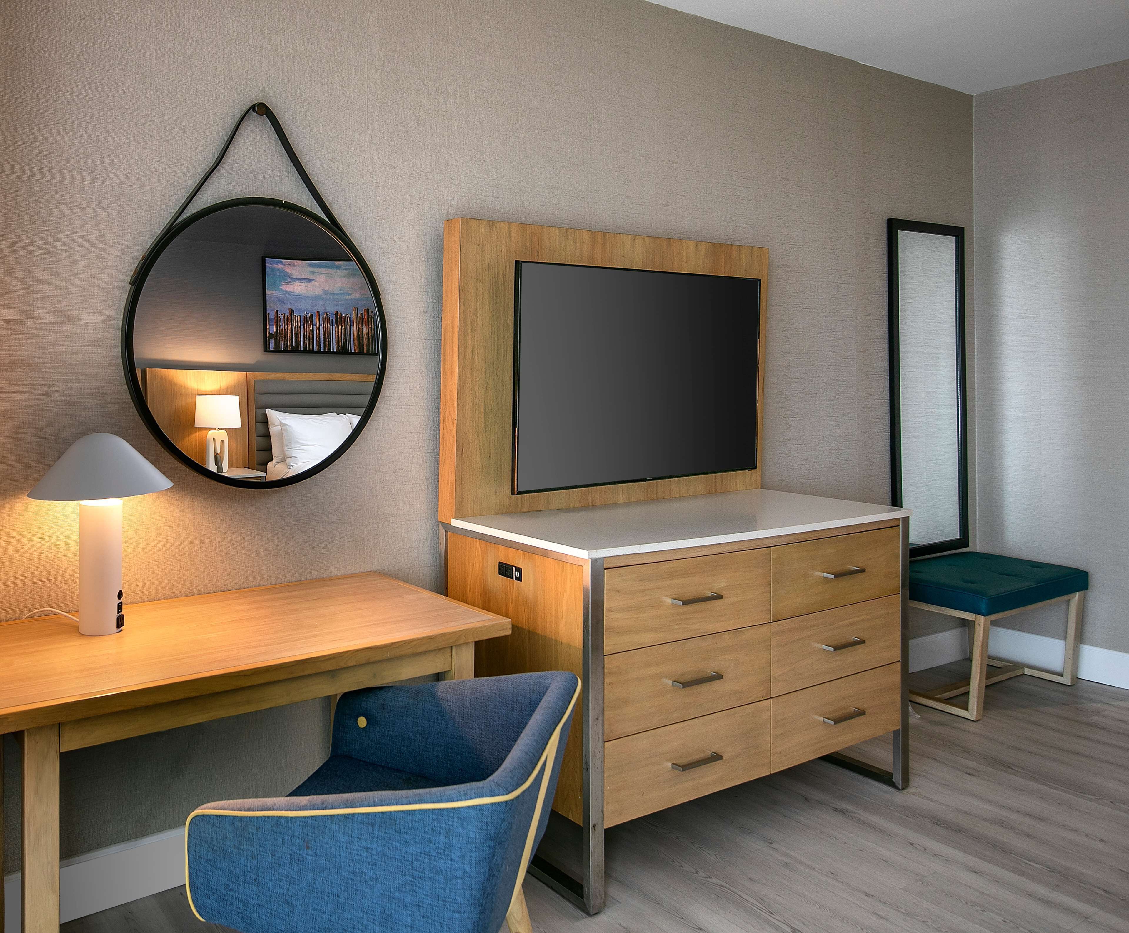 Une chambre d’hôtel avec un bureau en bois, une chaise bleue, une télévision, un miroir et une table de chevet avec une lampe.