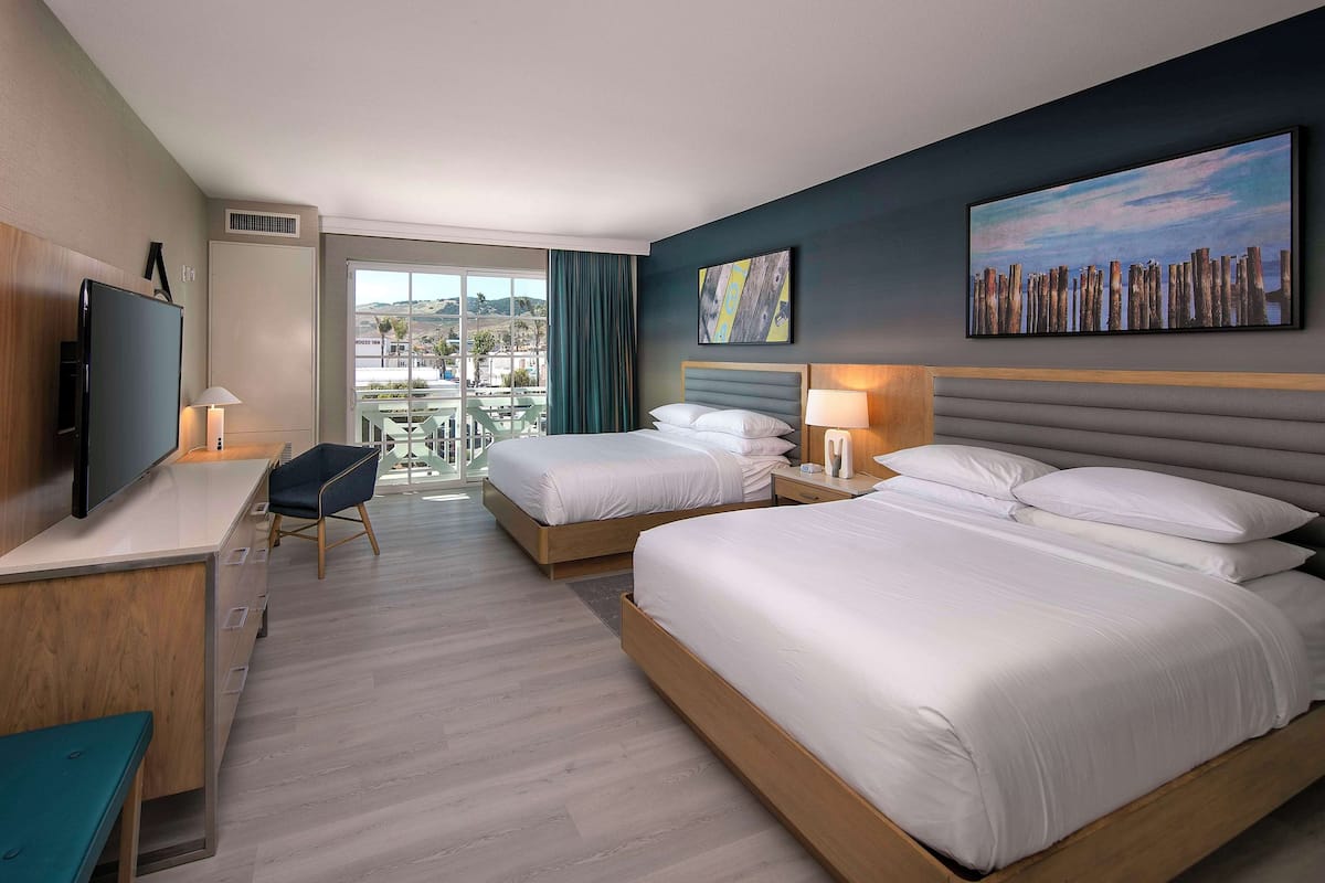 A hotel room with two beds, a desk, a TV, and a balcony with a view.