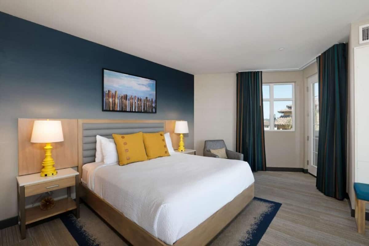 A hotel room with a bed, two bedside tables with lamps, a chair, a window with curtains, and a painting on the wall.