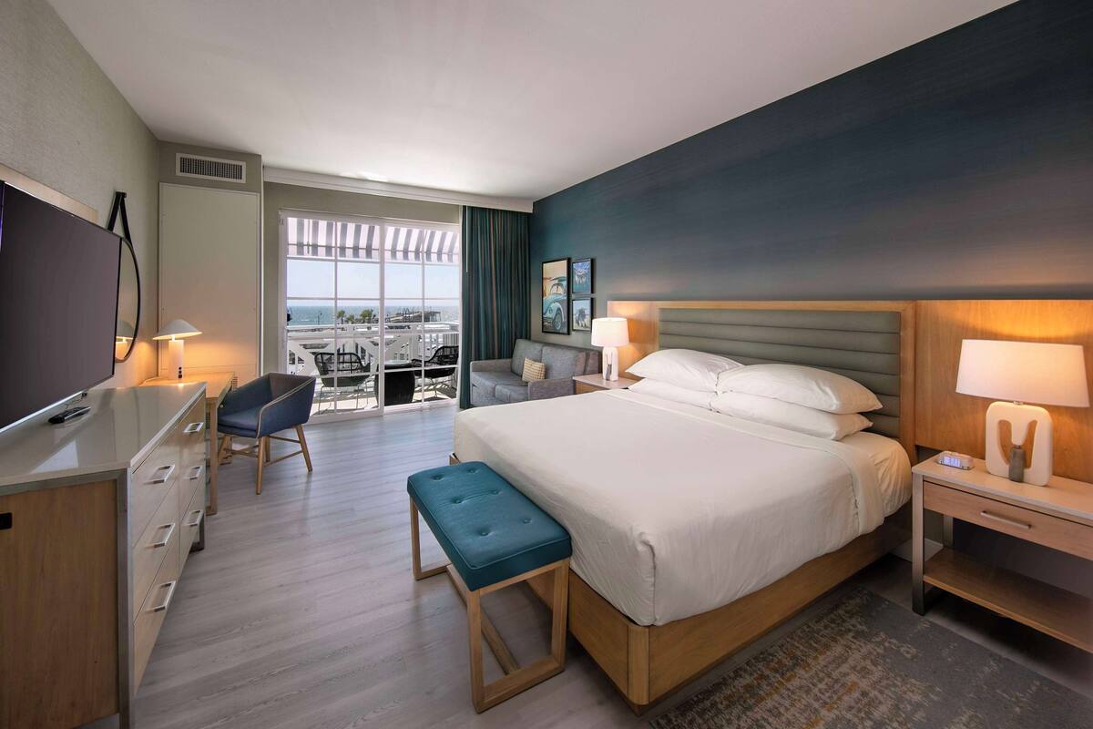 A hotel room with a bed, a desk, a TV, and a balcony with a view.