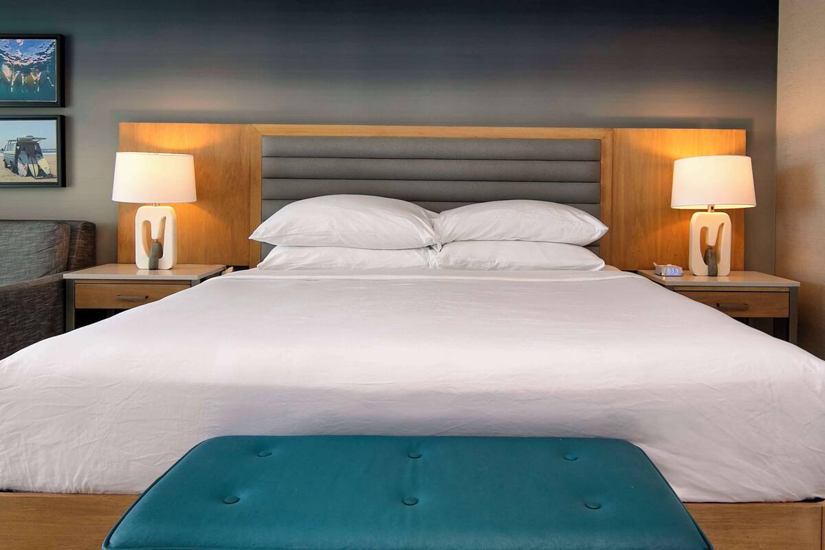 A neatly made bed with a teal bench in front, flanked by two bedside tables with lamps, in a room with a dark ceiling and framed pictures on the wall.