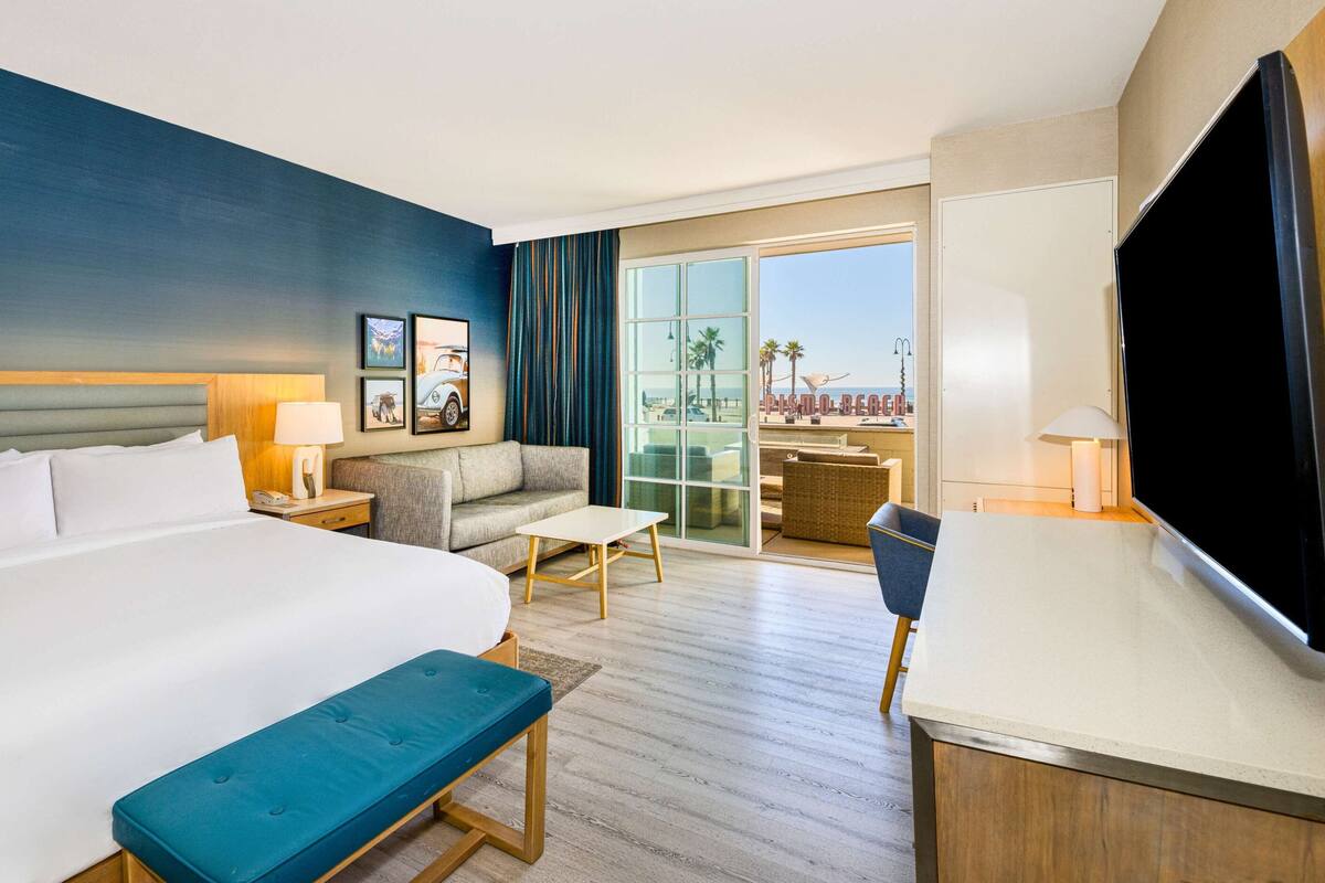 A hotel room with a large bed, a sofa, a small table, a bench, a TV, and a balcony with a view of the beach.