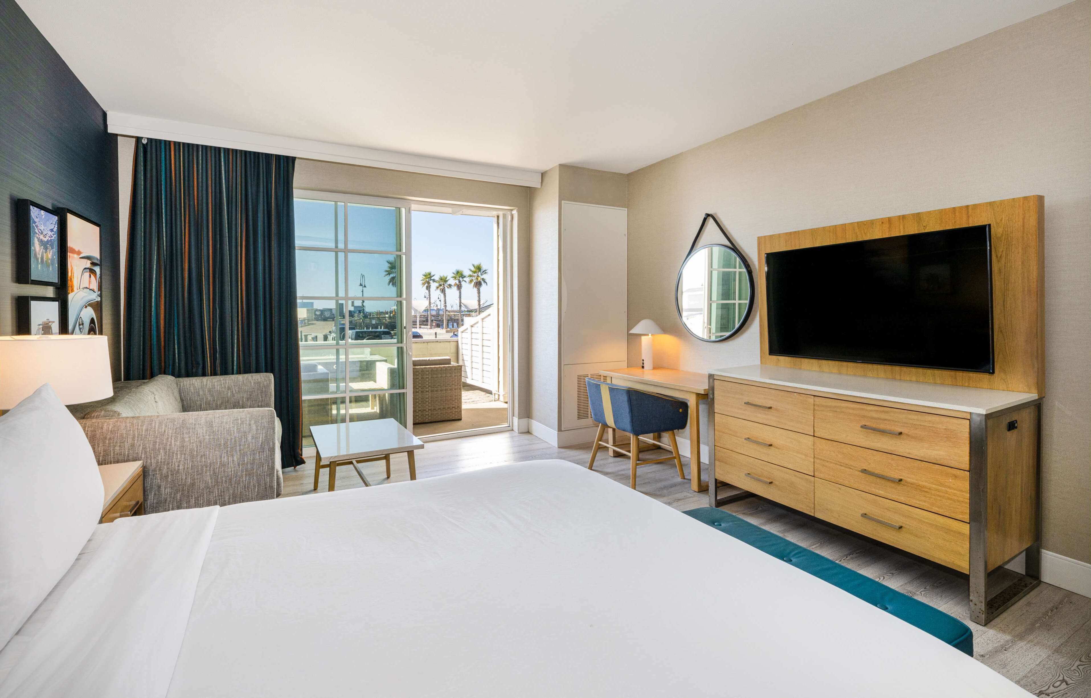 A hotel room with a large bed, a desk with a chair, a TV, a balcony with a view of palm trees, and a window with curtains.