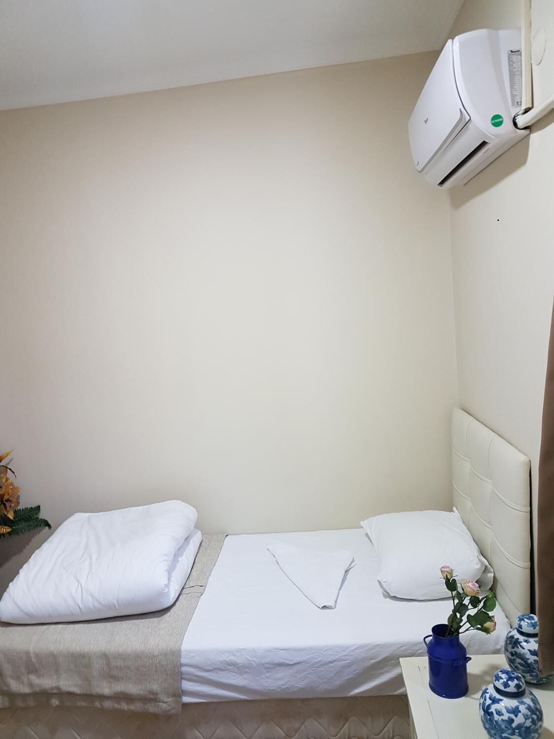 A small, neatly made bed with white linens and pillows, a blue vase, and a decorative plant on a bedside table.