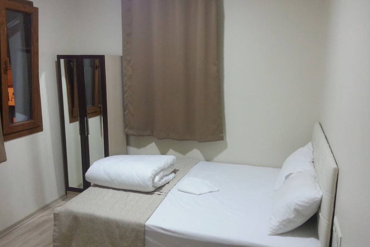 A small, clean room with a bed, a bench, a mirror, and a window with curtains.