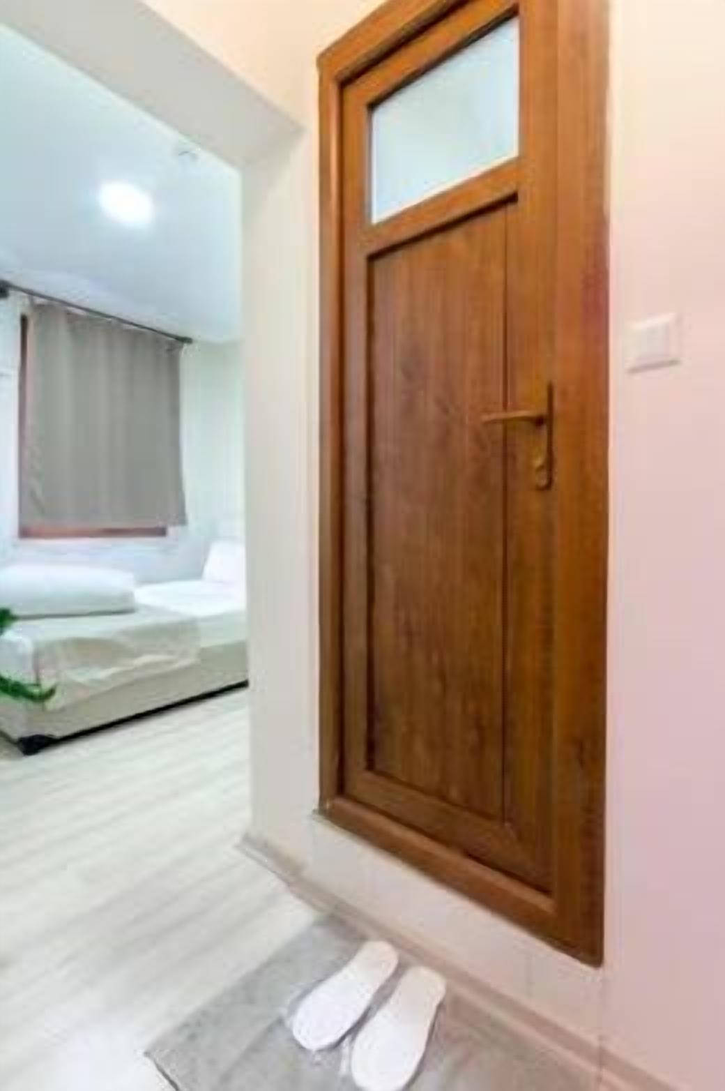 Una puerta de madera con un panel de vidrio, una cama con colchón blanco y manta gris, y piso de madera.