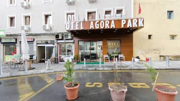Hotel llamado "Hotel Agora Park" con fachada moderna, zona de estar al aire libre y plantas en macetas en primer plano.